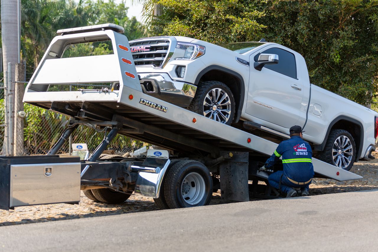 hero-img-01 Tow truck operator loading white GMC pickup truck on street in daytime.