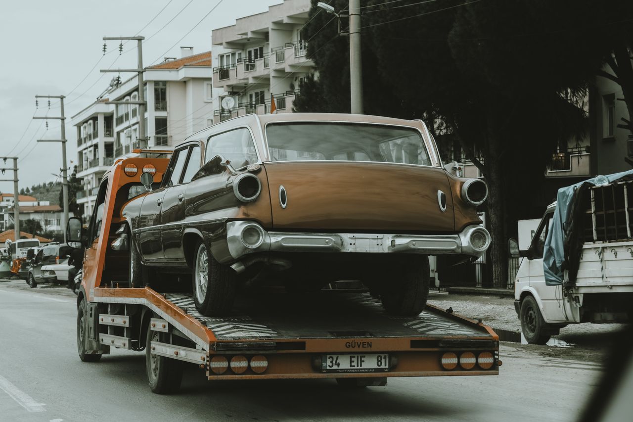 services-02 A vintage car loaded on a tow truck in a city street, surrounded by modern buildings.
